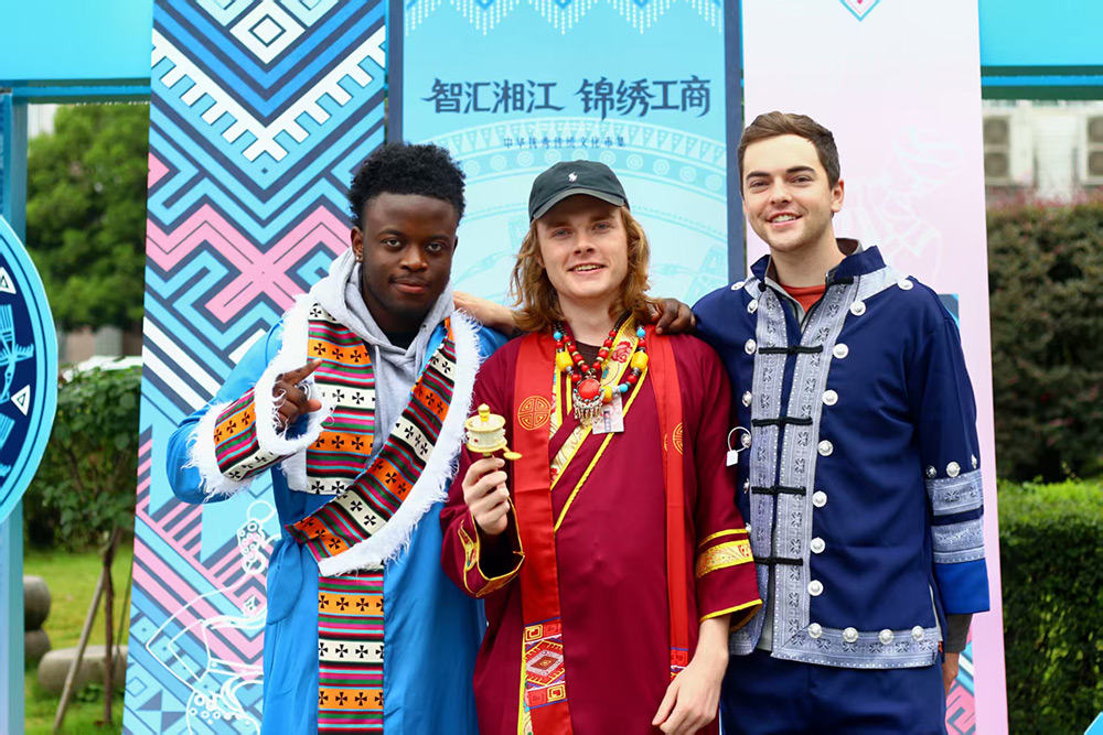 Three young men wearing colorful traditional-style clothing pose together and smile in front of a patterned cultural display backdrop
